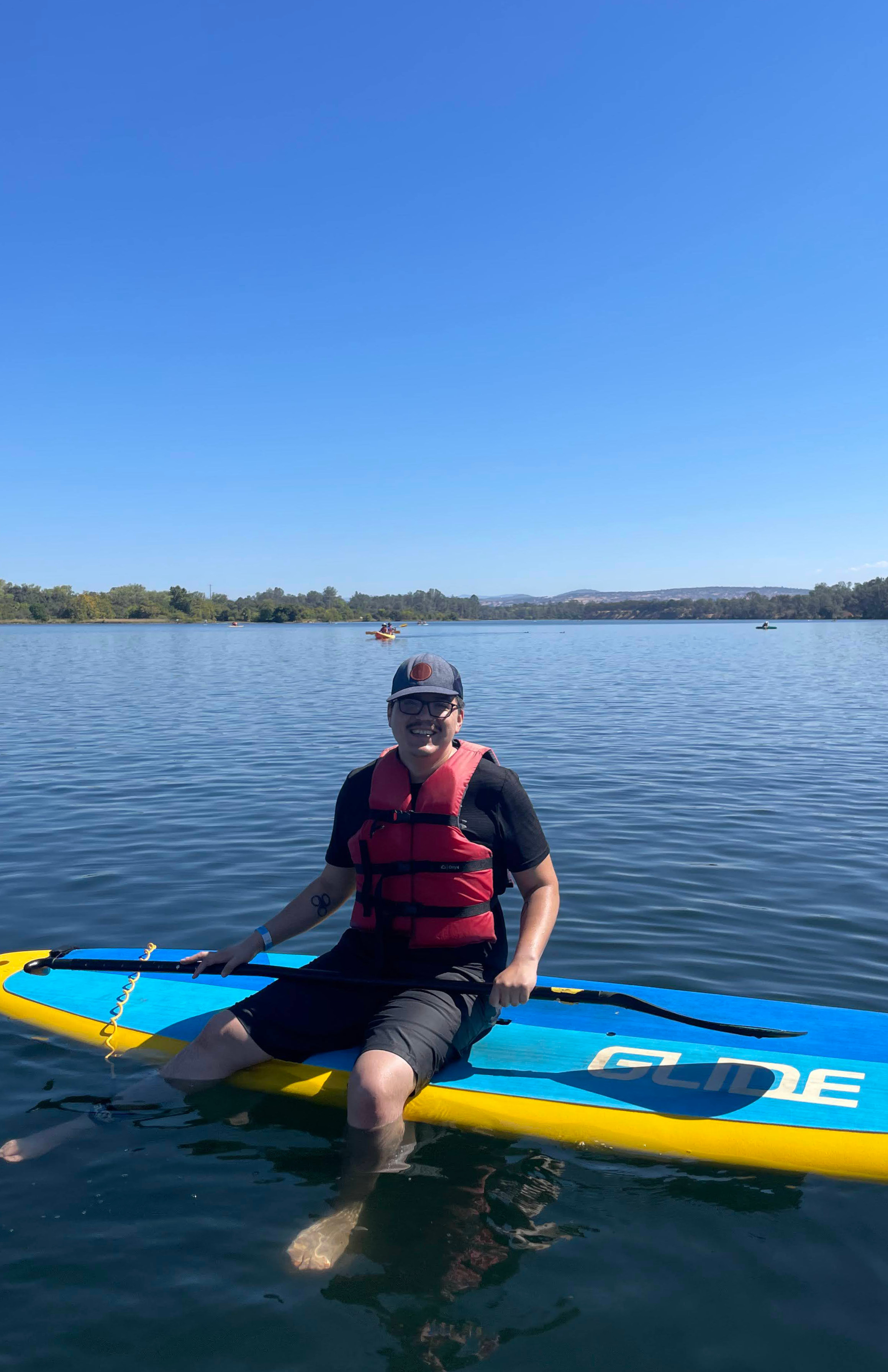 Man wearing a baseball cap and red floatation device sitting on a paddleboard